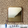 A mirror framed in wood hangs on a leopard print wall, with a vase of purple flowers and marble countertop below.