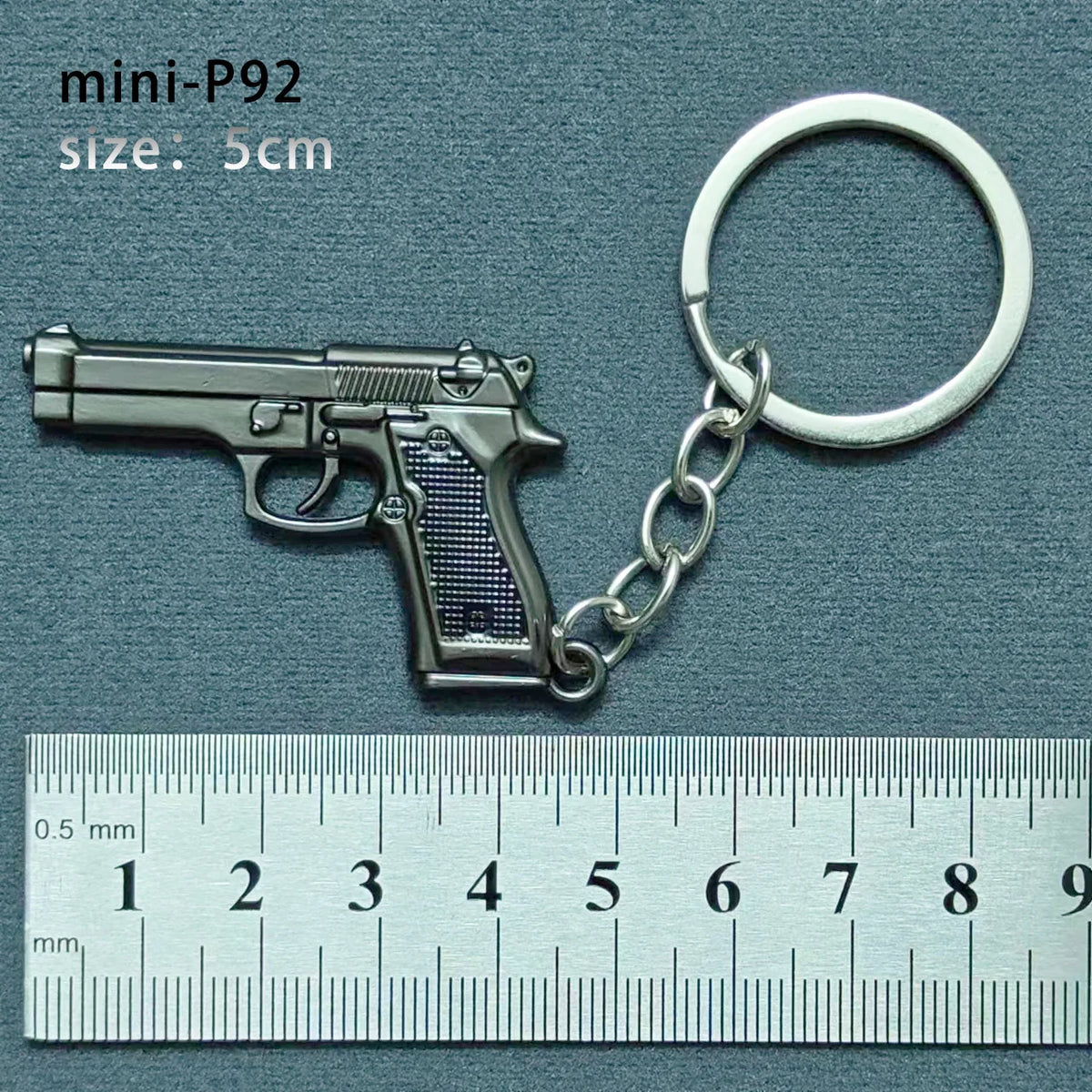 A silver miniature replica of a handgun is attached to a metal keyring, measured against a ruler.