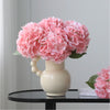 A bouquet of pink artificial hydrangea flowers in a white ceramic vase on a black table.