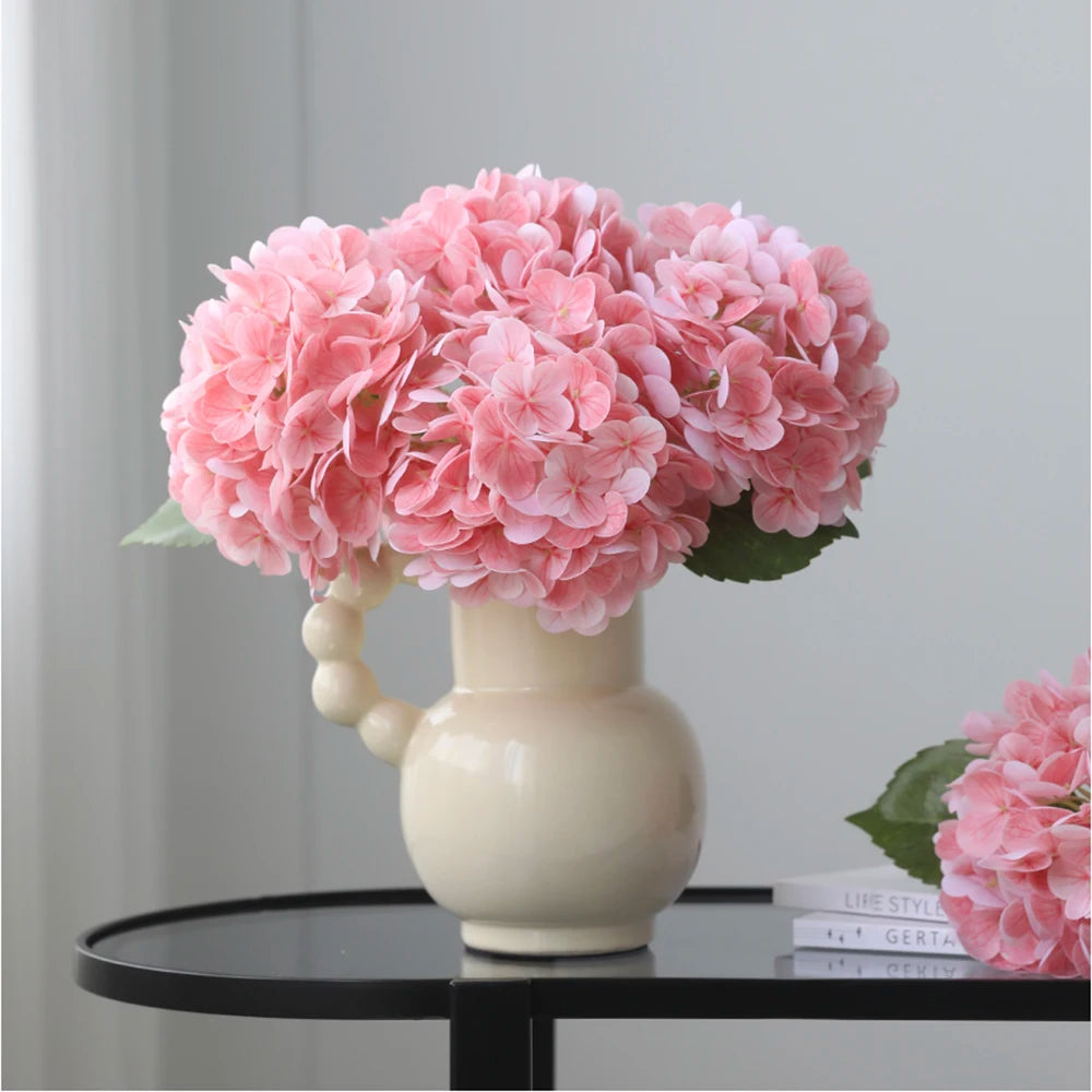 A bouquet of pink artificial hydrangea flowers in a white ceramic vase on a black table.