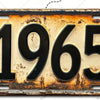 Primer plano de un cartel de pared de aluminio vintage con el año 1965 estampado, mostrando signos de desgaste y pátina.