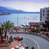 A red race car is driving on a curved asphalt track in front of a large building and blue sea.