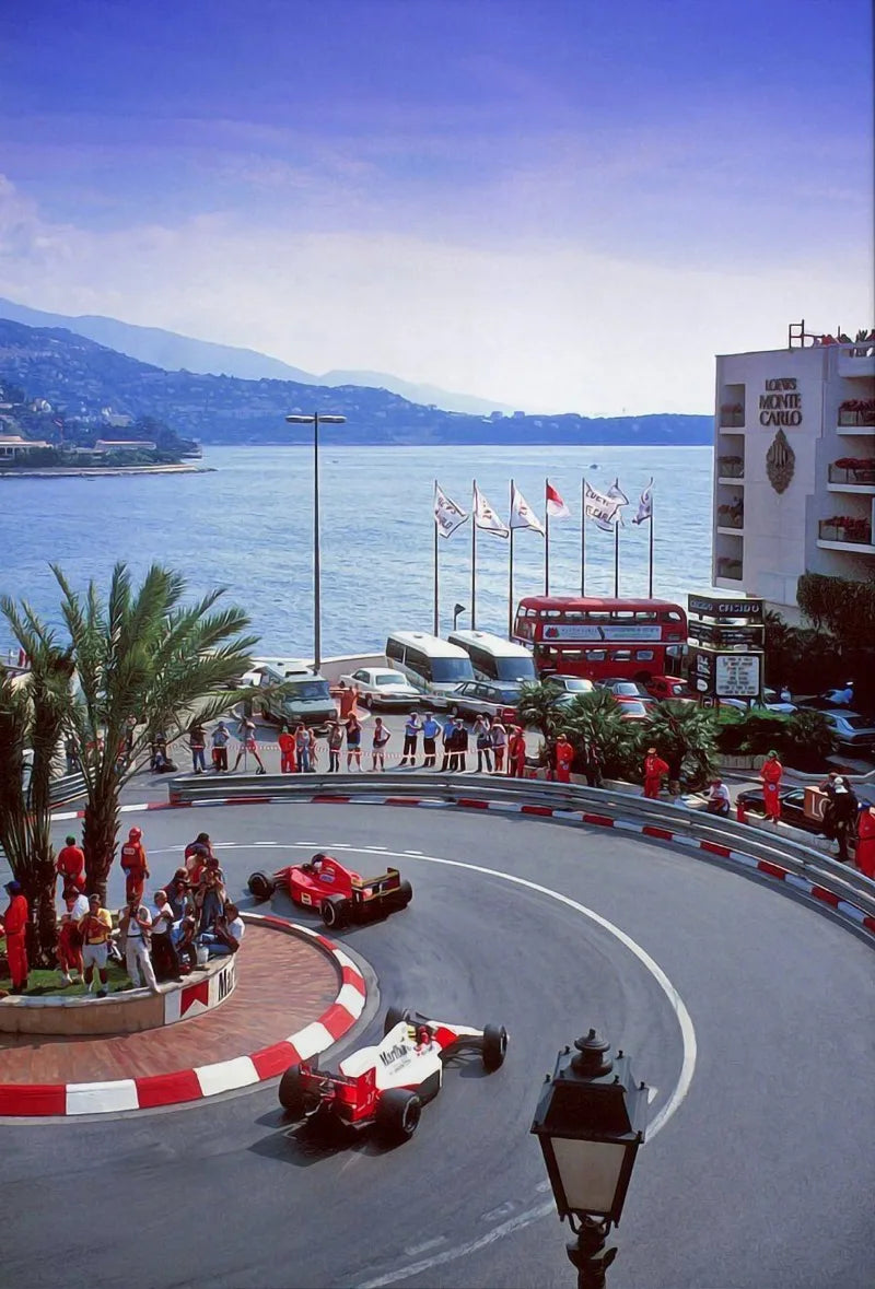 A red race car is driving on a curved asphalt track in front of a large building and blue sea.