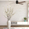 A vase holds a branch of white faux plum blossoms against a white brick wall.