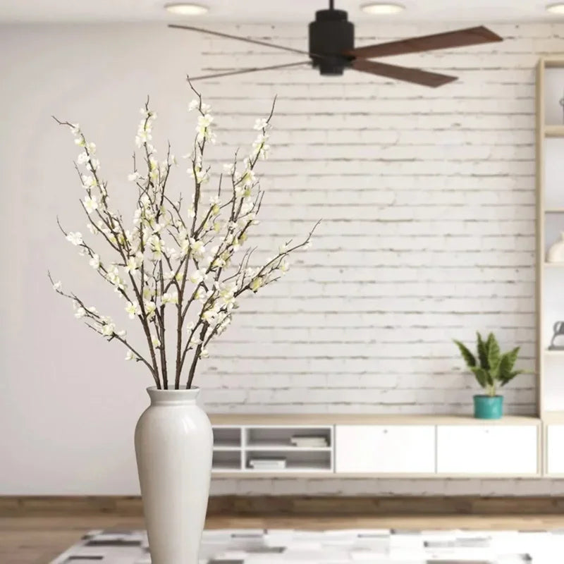 A vase holds a branch of white faux plum blossoms against a white brick wall.