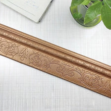 A close-up of a gold-colored self-adhesive 3D wall trim line featuring an ornate floral pattern.