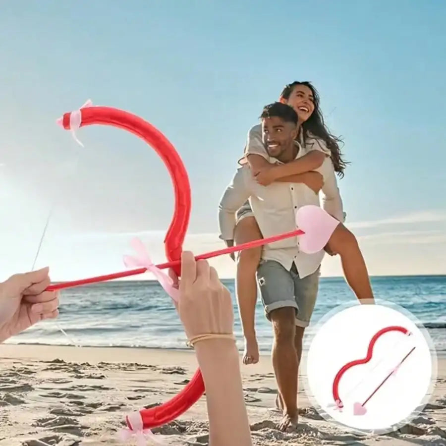 A couple is using a red Cupid heart bow and arrow prop on a beach with the ocean in the background.
