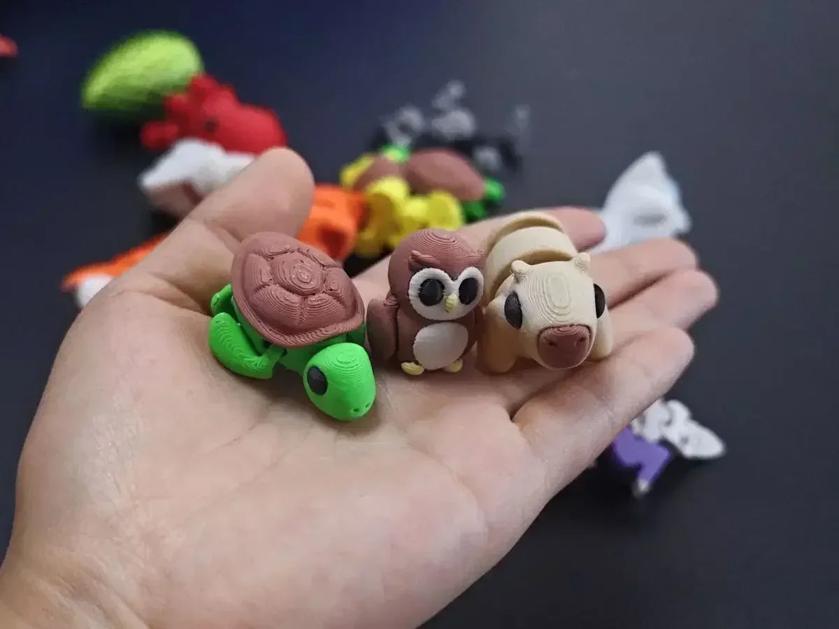 A hand holds several small, articulated 3D printed animal figurines including a green turtle, a brown owl, and a beige mammal