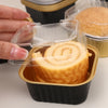 A hand is holding a square aluminum foil baking box with a transparent lid containing a rolled cake or pastry.