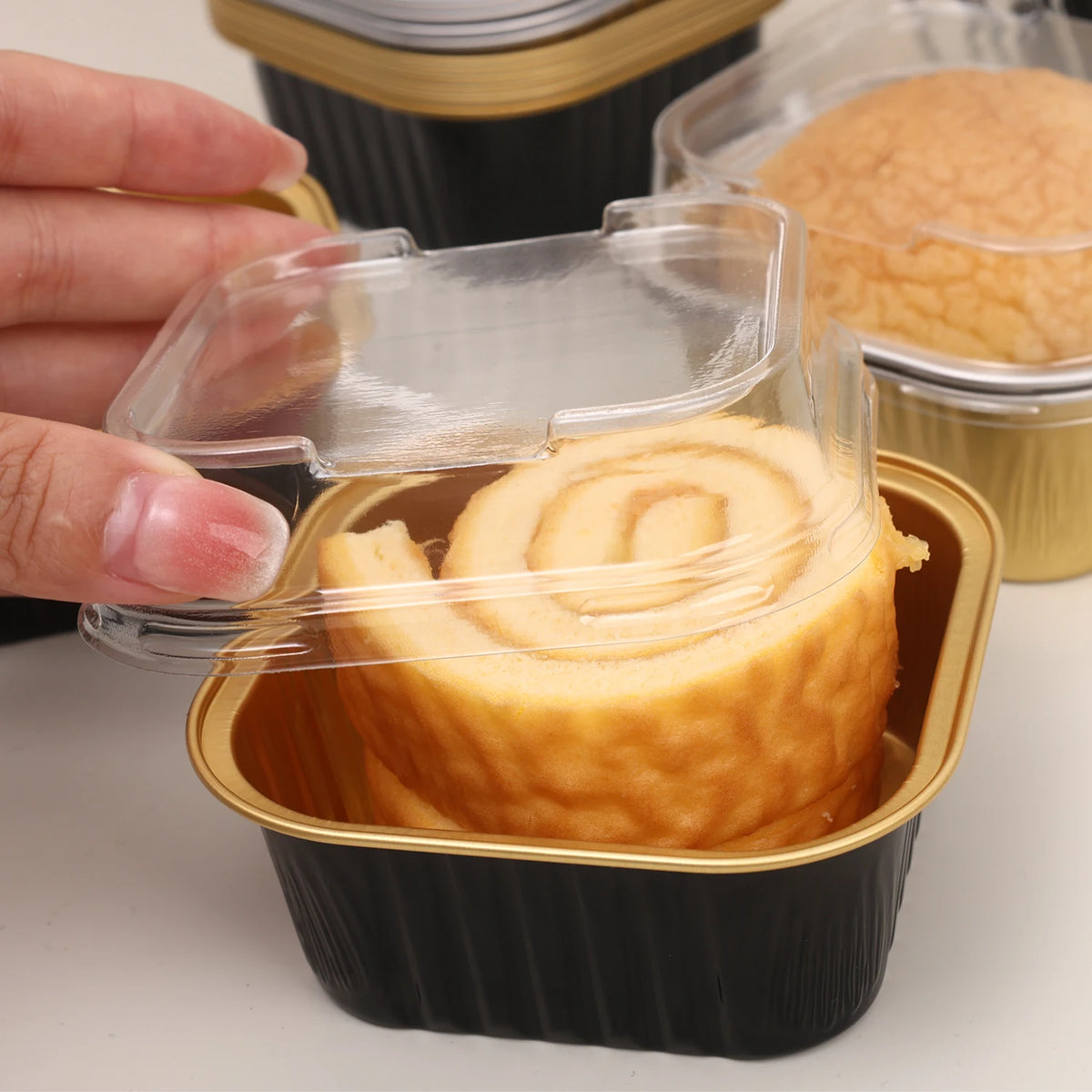 A hand is holding a square aluminum foil baking box with a transparent lid containing a rolled cake or pastry.