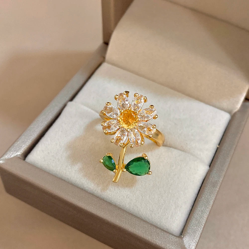 A gold cocktail ring featuring a yellow flower design with green leaves, displayed in a light-colored box.