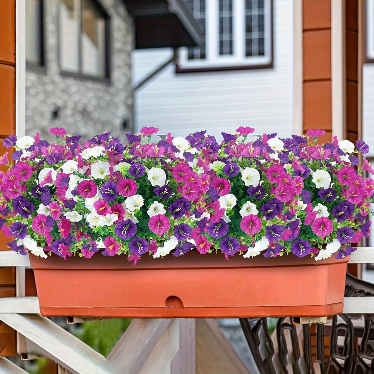 A vibrant mixed planting of purple, pink, and white artificial flowers in an orange planter on a wooden railing.