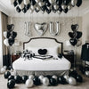 Un lit décoré de ballons en forme de cœur argentés et noirs, face à un mur de briques blanches.