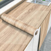 A roll of self-adhesive wood grain wallpaper is placed on a light wood countertop next to a sink.