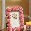 A pink rose resin picture frame decorated with artificial roses, resting on a wooden surface.