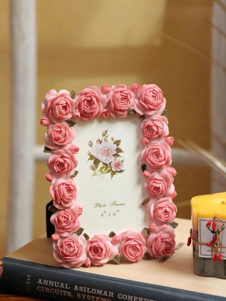 A pink rose resin picture frame decorated with artificial roses, resting on a wooden surface.