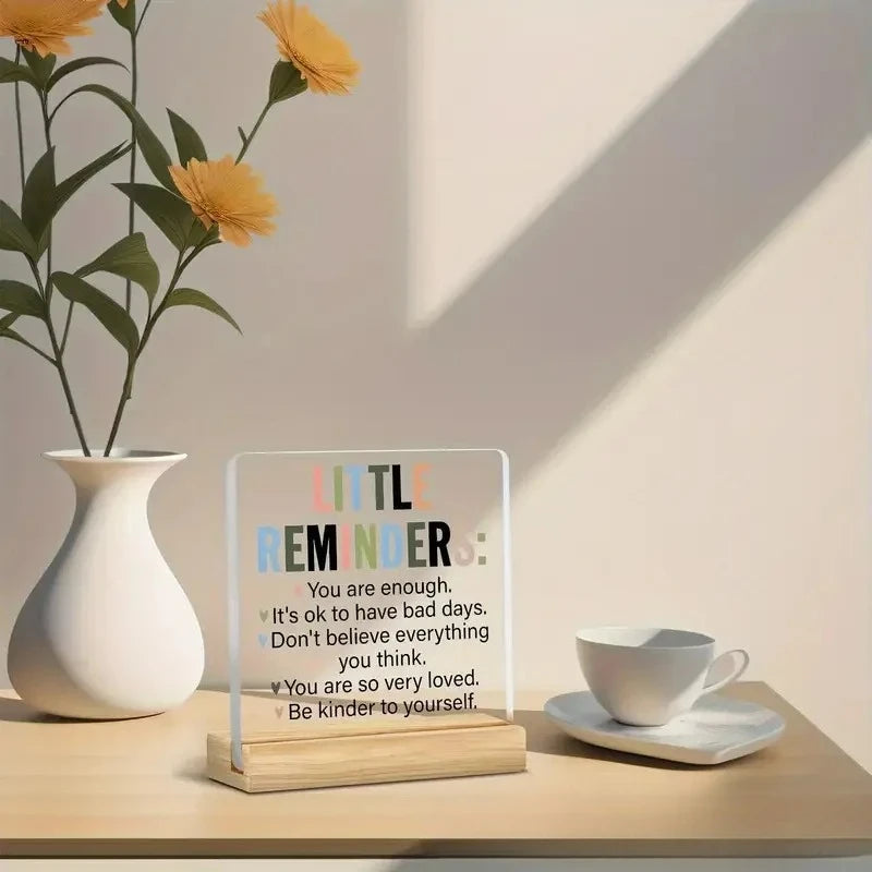 An acrylic desk sign with a positive message about self-worth sits on a wooden surface next to a white cup and a plant.