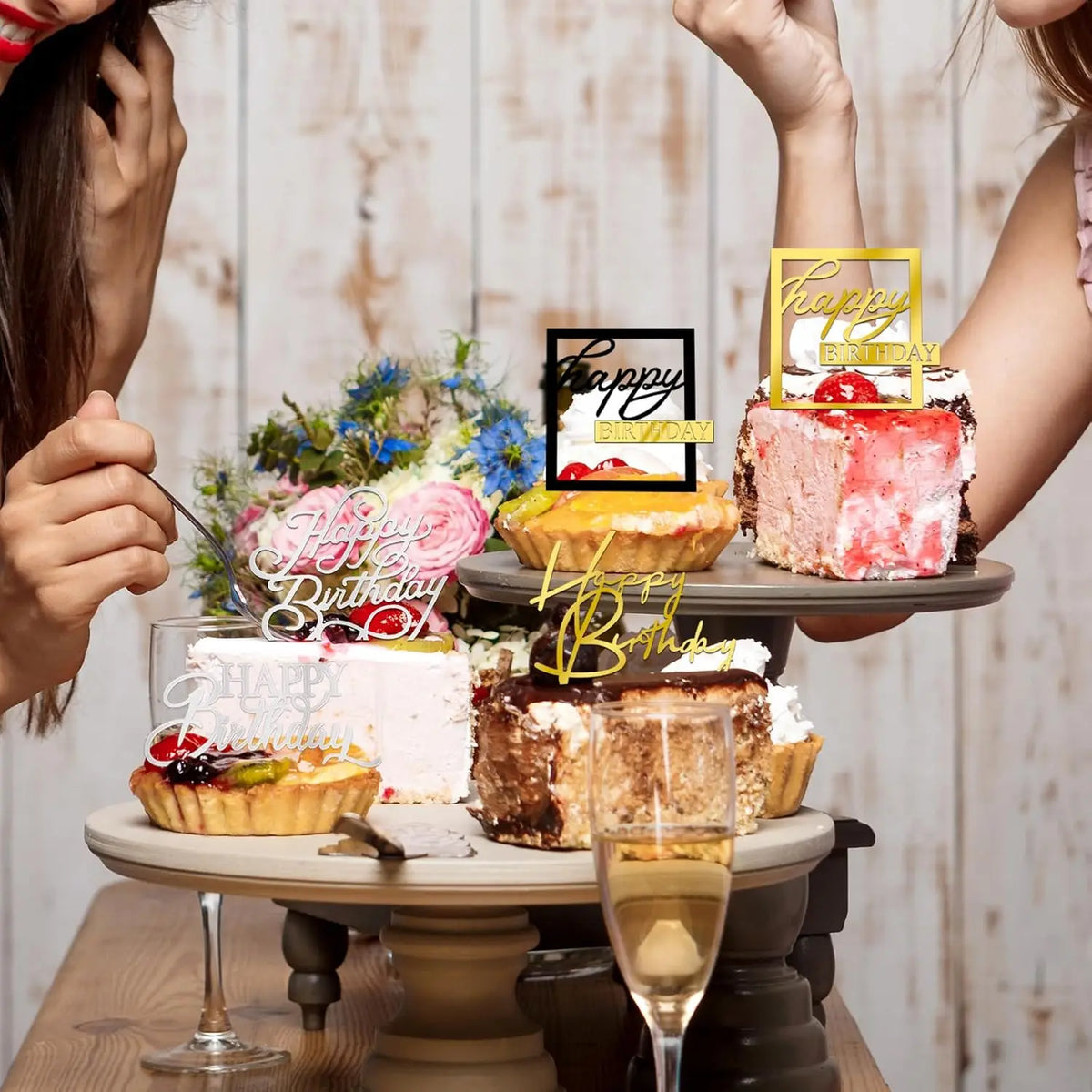 A tiered display of birthday cakes and mirrored gold acrylic cake toppers being held by two people.
