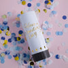 A white tube of Hand Twist Confetti Cannons lying on pink surface surrounded by scattered confetti.