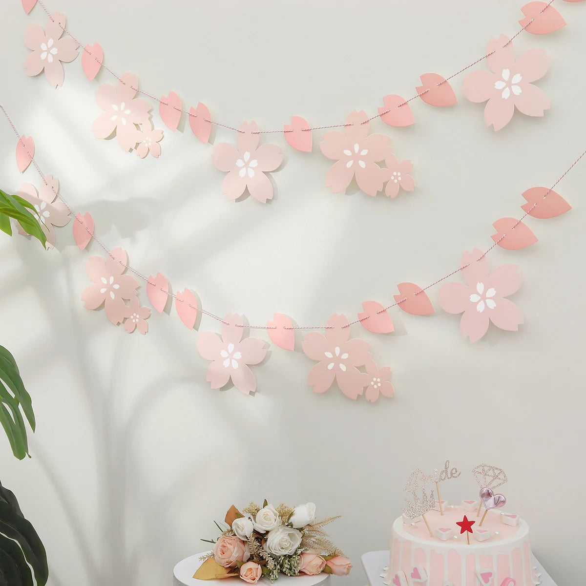 Pink cherry blossom paper flag banner strung across a white wall with soft shadows.