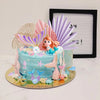A mermaid-themed cake decorated with blue frosting, pink and purple fins, shells, and sea creatures, set against a black fram