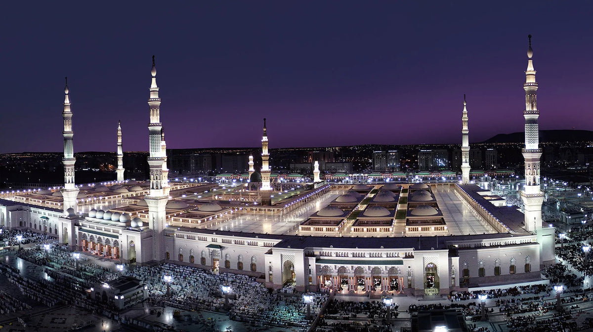 A panoramic view of the Masjid an Nabawi, featuring minarets and architecture, illuminated at night.