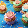 A variety of green and pink frosted cupcakes decorated with happy birthday toppers on a blue wooden surface.
