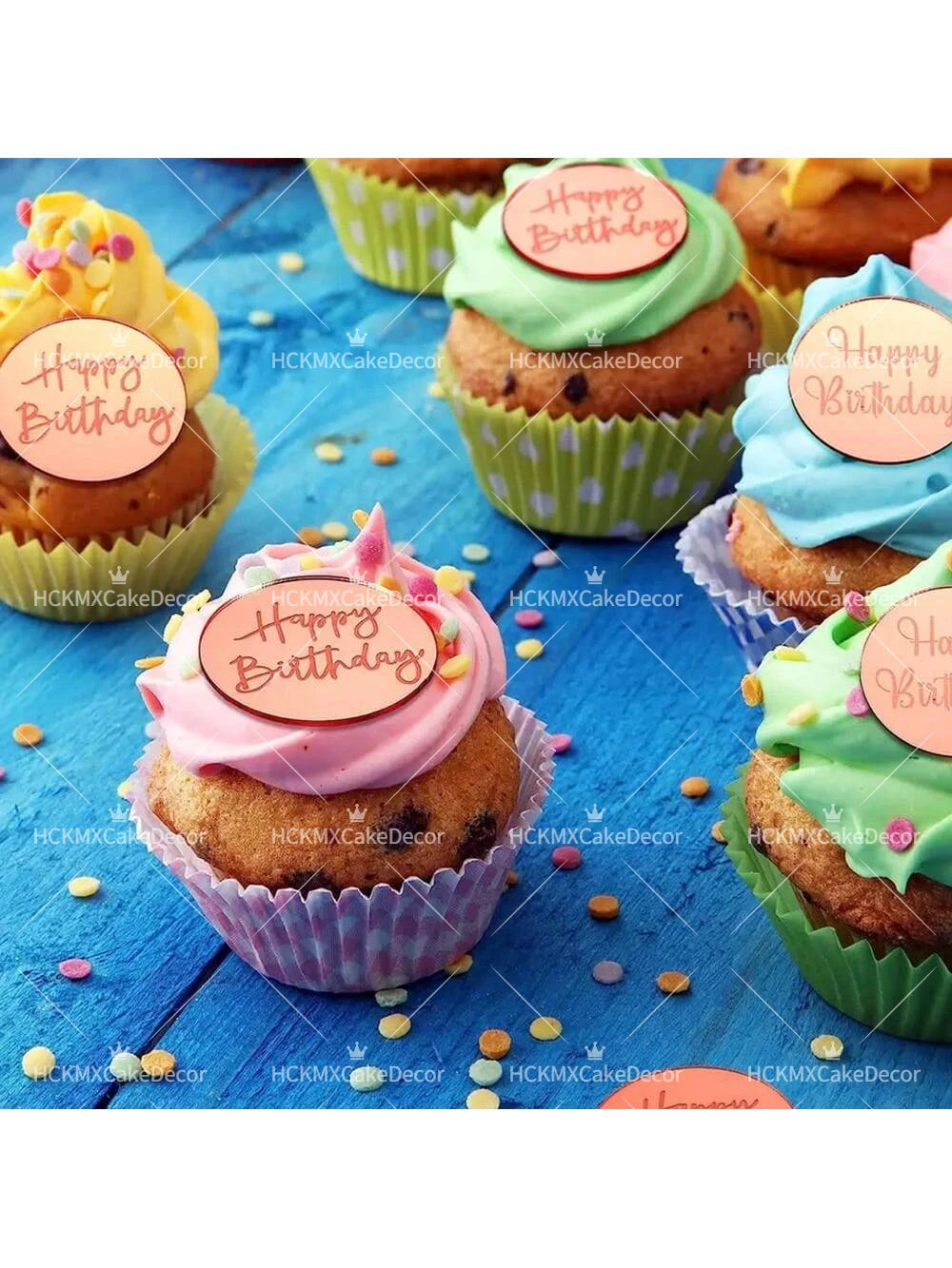 A variety of green and pink frosted cupcakes decorated with happy birthday toppers on a blue wooden surface.