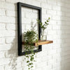 A black wooden floating wall shelf displaying a glass vase with greenery on a white brick wall.