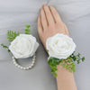 A hand is holding a white rose corsage bracelet with green foliage on a gray fabric background.