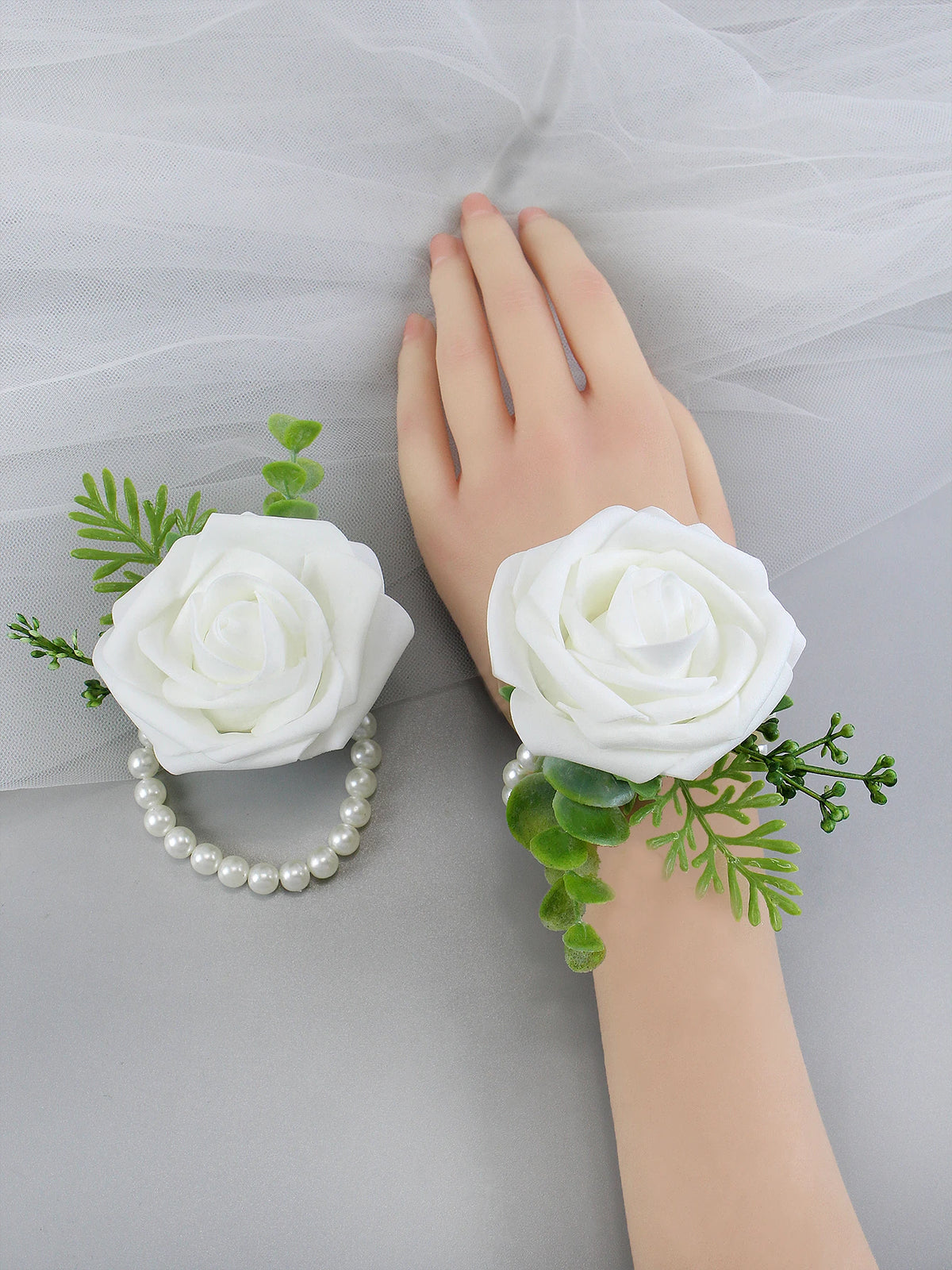 A hand is holding a white rose corsage bracelet with green foliage on a gray fabric background.