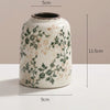 A white ceramic vase with green and brown floral patterns, shown against a neutral background.
