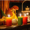 A collection of four pillar candles in orange, green, red, and yellow glow on a wooden surface with mushrooms in the backgrou