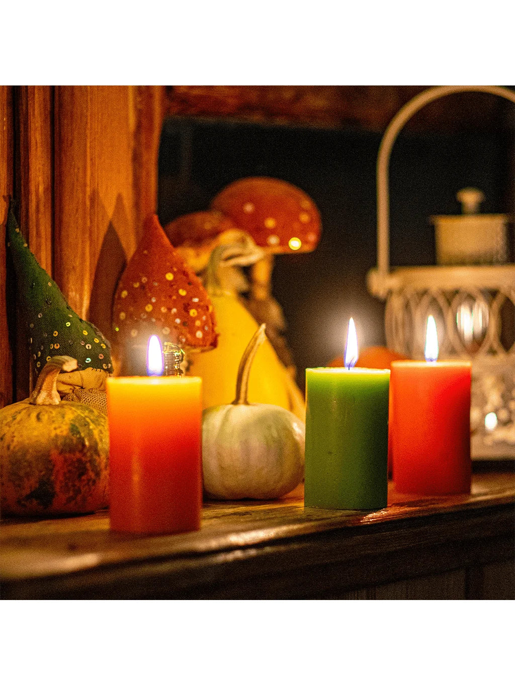 A collection of four pillar candles in orange, green, red, and yellow glow on a wooden surface with mushrooms in the backgrou