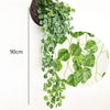 Une longue cascade de plantes d'ivy vertes artificielles suspendues dans un panier en rotin foncé sur fond blanc.