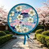 A blue floral foldable pocket fan is displayed outdoors in a garden setting with pink cherry blossoms and green shrubs.