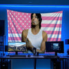 A man sits in front of a large American flag banner decorated with pink and white stripes and stars, with computer monitors v