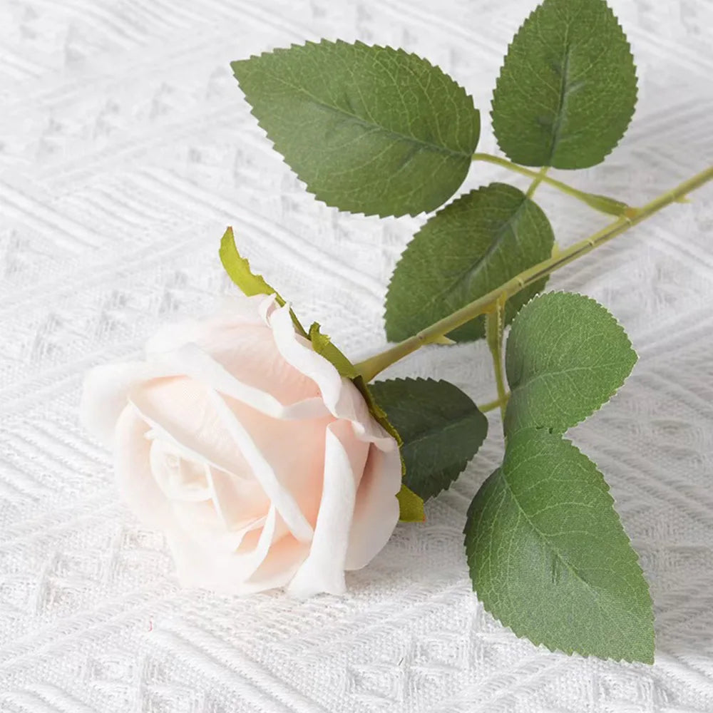 A soft pink artificial rose with green leaves on a white textured background.