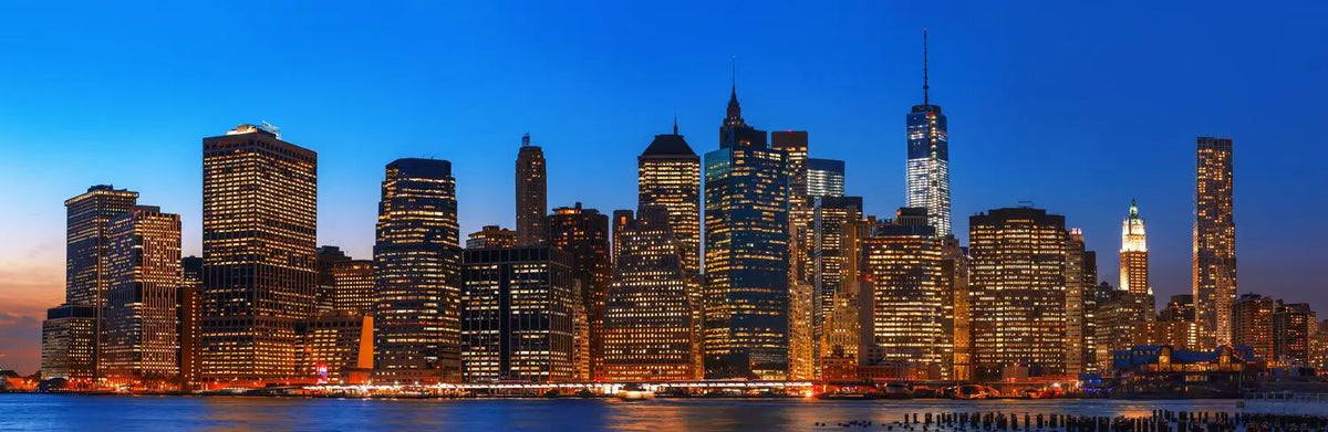 A nighttime view of the New York City skyline featuring illuminated skyscrapers against a deep blue sky.