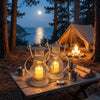 A decorative metal candlestick lantern with a lit candle sits on a wooden table in a forest setting at twilight.