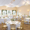 Elegant dining setup with white stretch tablecloths, gold chairs, and sheer white drapery in a bright room.