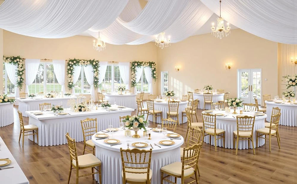 Elegant dining setup with white stretch tablecloths, gold chairs, and sheer white drapery in a bright room.