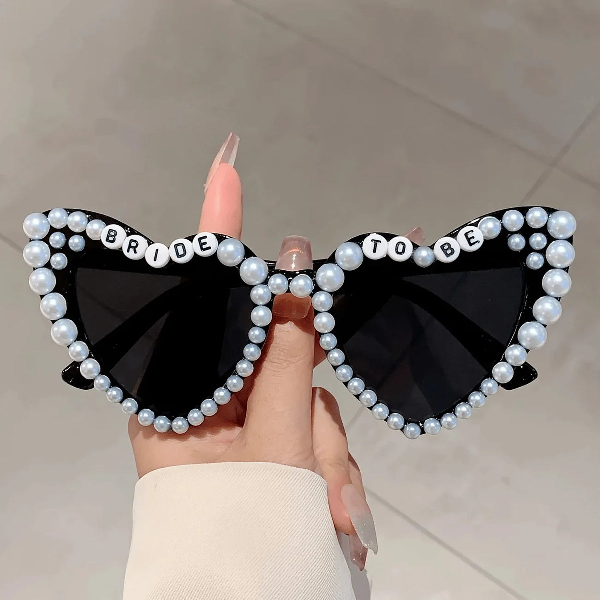 A hand holds a pair of black heart-shaped glasses decorated with white pearls and the words bride to be
