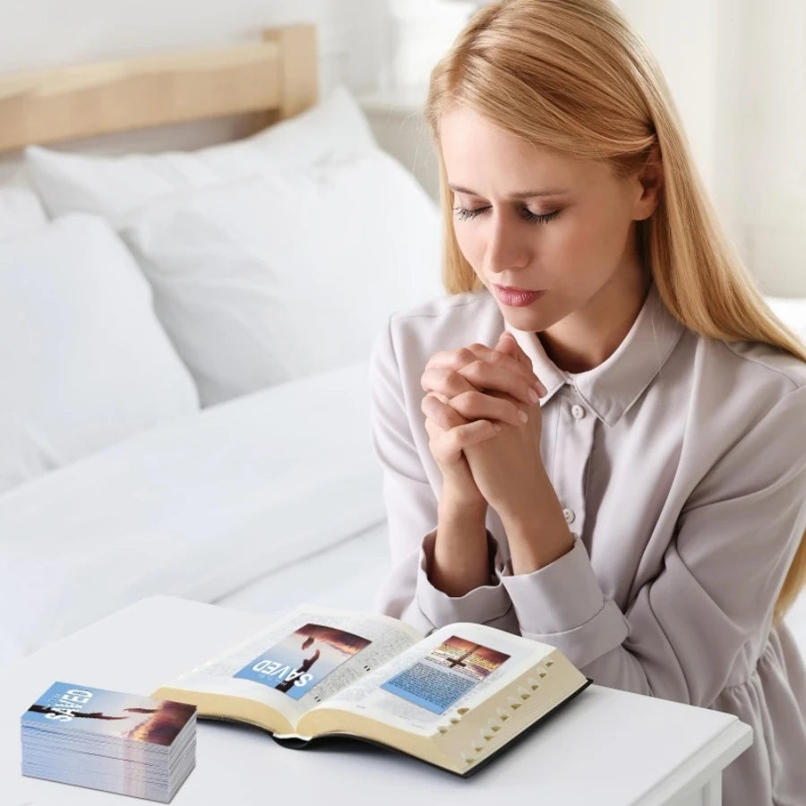A blonde woman is praying with her hands over an open inspirational prayer card book on a white table.