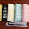 A row of pink, green, yellow, and black 100 envelope savings challenge binders displayed on a wooden surface.