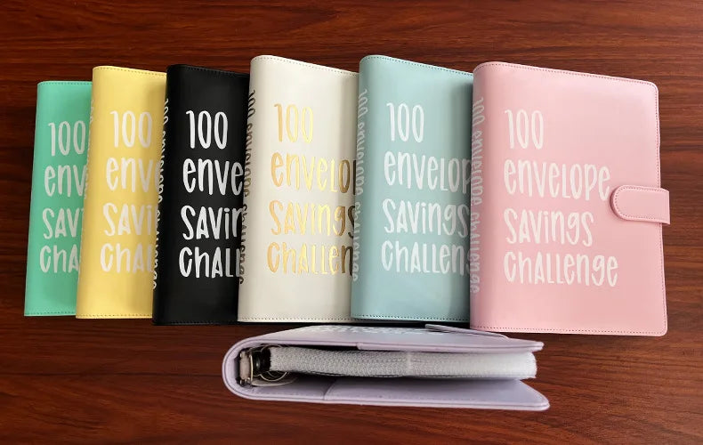 A row of pink, green, yellow, and black 100 envelope savings challenge binders displayed on a wooden surface.
