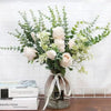 A bouquet of artificial eucalyptus stems with white roses and green foliage in a glass vase.