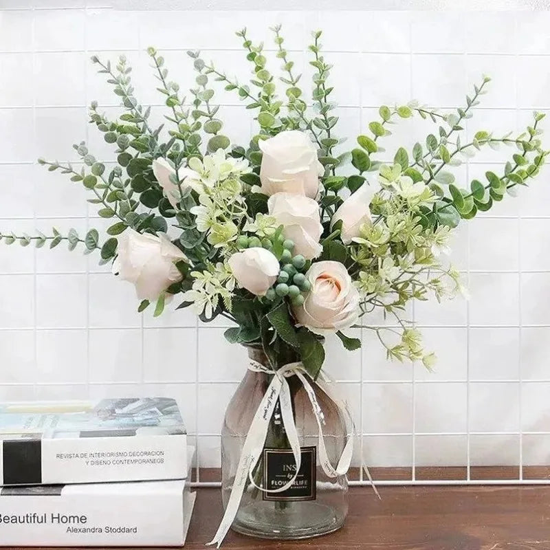 A bouquet of artificial eucalyptus stems with white roses and green foliage in a glass vase.