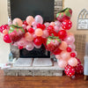 A balloon arch display featuring pink, red, and green strawberry-shaped balloons decorating a fireplace mantel.