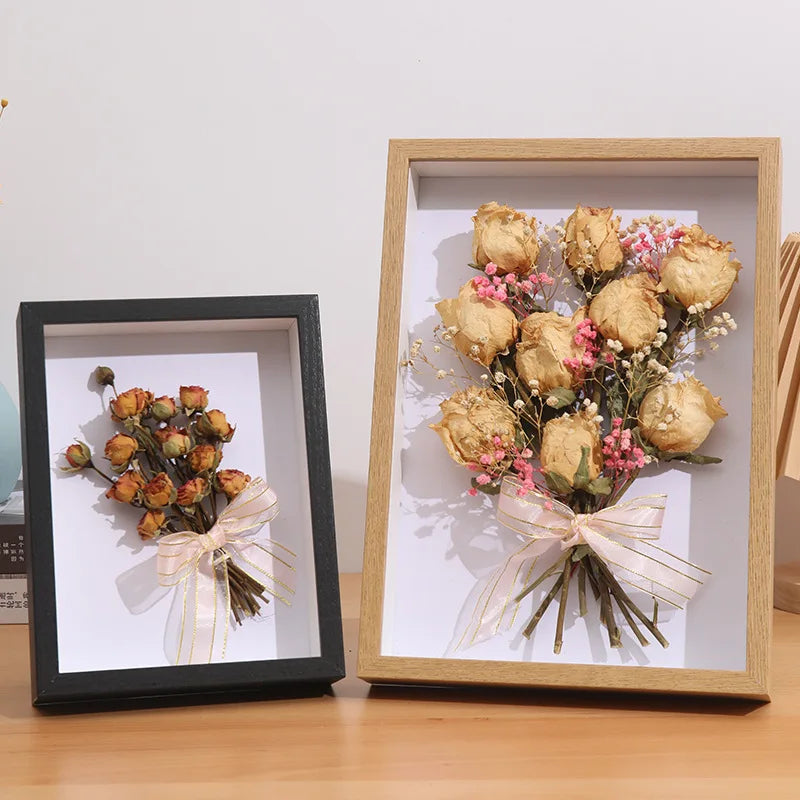 A wooden shadow box frame containing dried floral arrangements tied with sheer ribbon and natural greenery.